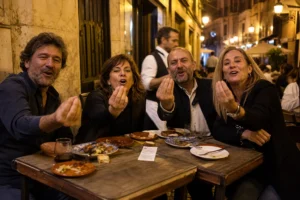 Cuatro comensales adultos en una terraza de Madrid alzan a la vez la mano derecha gesticulando «¡vamos!», sin mirar el ticket que el camarero ha dejado en el centro de la mesa, calle Cava Baja iluminada por farolas amarillas