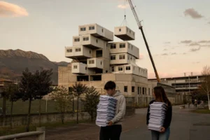 Vista general de un campus de Formación Profesional vasco al atardecer con una grúa colocando varios módulos blancos prefabricados encima del edificio existente, dándole al conjunto un aspecto inestable como de bloques apilados; en primer plano dos estudiantes con papeles caminan confusos