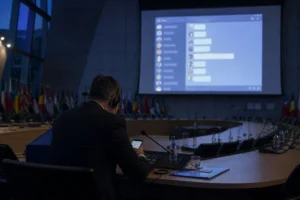 Hemiciclo de la OTAN en penumbra con un único delegado tecleando en su móvil ante una gran pantalla que proyecta un grupo de WhatsApp con muchos avatares de diplomáticos